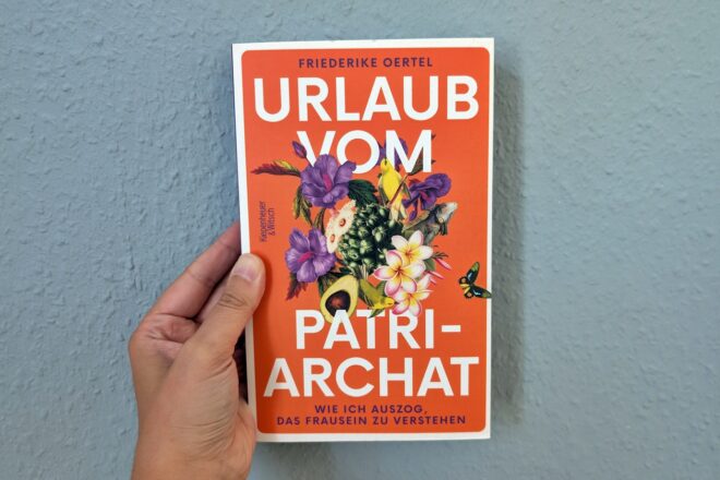 Eine Hand hält das Buch "Urlaub vom Patriarchat" von Friederike Oertel gegen eine hellblaue Wand.