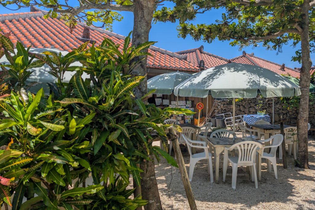 Café am Ortseingang von Taketomi. Im Vordergrund sieht man eine Pflanze, dahinter sind Plastikstühle und Schirme und im Hintergrund kann man die roten Schieferdächer erahnen.