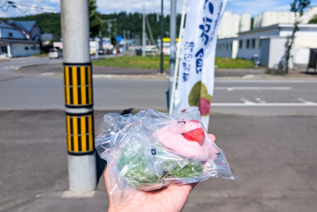 Ein Hand hält Mochis in die Kamera. Im Hintergrund ist eine Straßenkreuzung in Biei zu sehen.