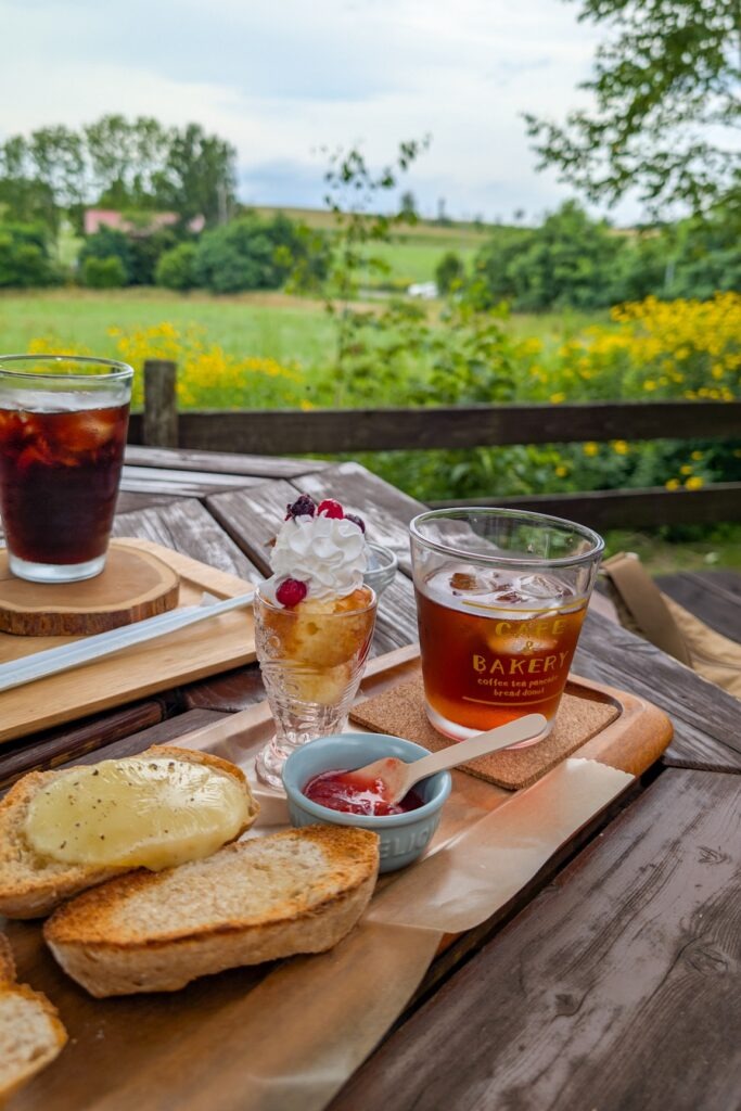 Tablett mit Baguette, Marmelade, Kuchen, Eiskaffee und -tee. Im Hintergrund sieht man ein Feld. Auf diesem blühen die für Hokkaido im Sommer typischen Blumen.