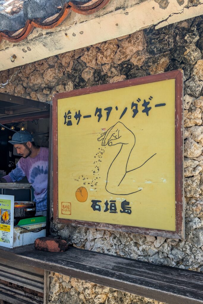 Schild eines Imbiss am Aussichtspunkt zur Kabira Bay. Man sieht eine Hand, welche einen orangenen, kreisförmigen Gegenstand würzt. Im Hintergrund steht ein Mitarbeiter des Imbiss.