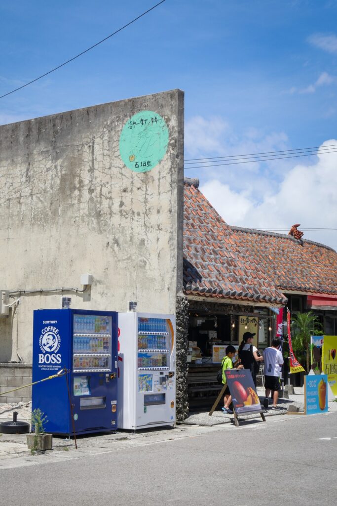 Zwei Vending Machines stehen an einer grauen Mauer. Auf der rechten Seite der Mauer ist ein Verkaufsstand in einem Haus, an welchem gerade eine dreiköpfige Familie vorbei geht.