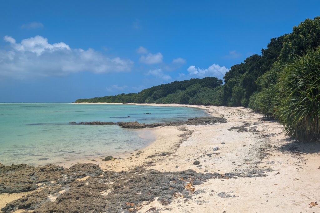 Strand auf Taketomi. Links ist das türkisfarbene Wasser, dazwischen ein schmaler Streifen Sandstrand und auf der rechten Seite dichte, grüne Vegetation.