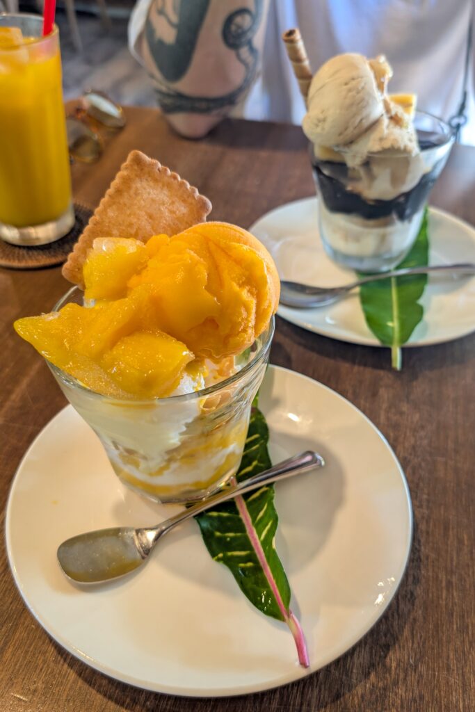 Im Vordergrund sieht man einen Mangoeisbecher und im Hintergrund einen mit Kaffee. Daneben steht noch ein Saft und ein Mensch stützt sich mit dem Ellenbogen auf dem Tisch ab.