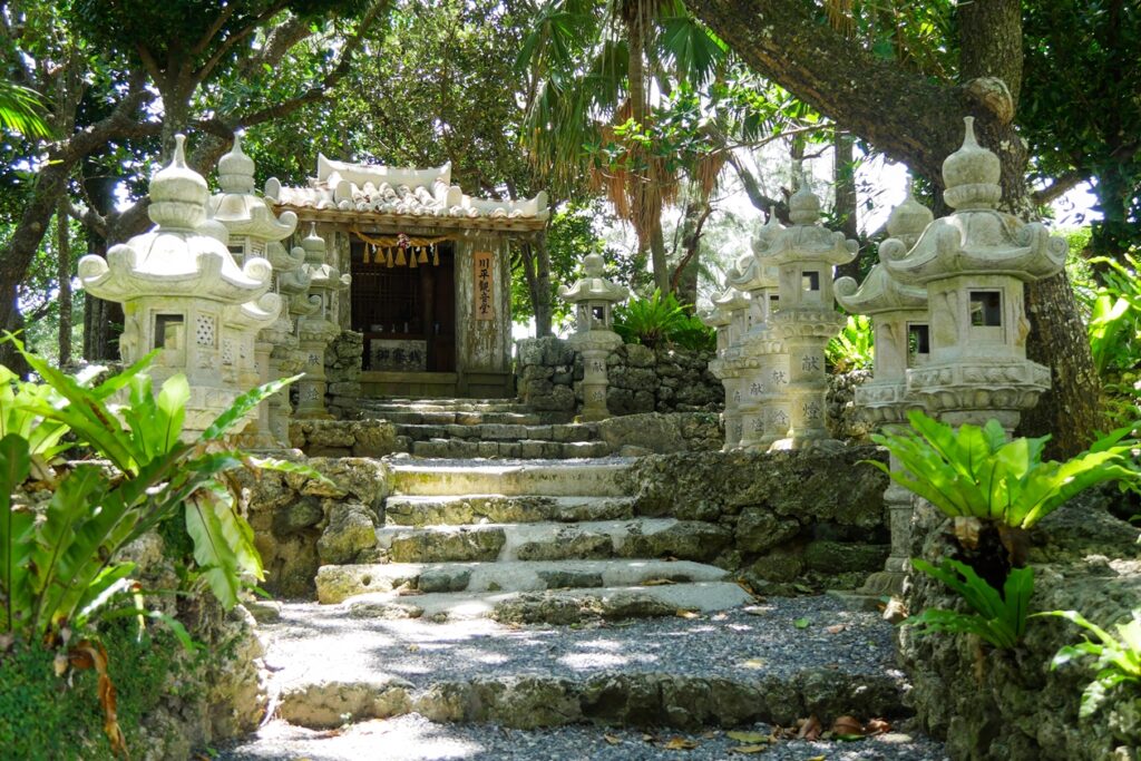 Schrein in der Nähe des Aussichtspunkt auf die Kabira Bay. Eine Treppe führt zu ihm hoch, welche links und rechts von grünen Pflanzen und kleinen Staturen gesäumt ist.