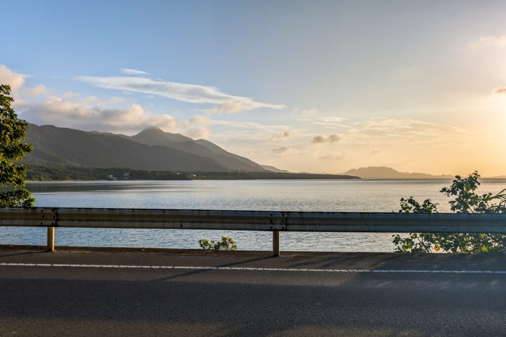Sonnenuntergangsstimmung am Rande der Straße auf Ishigaki. Der Blcik fällt auf die Leitplanke, das Meer dahinter und einige Berge.
