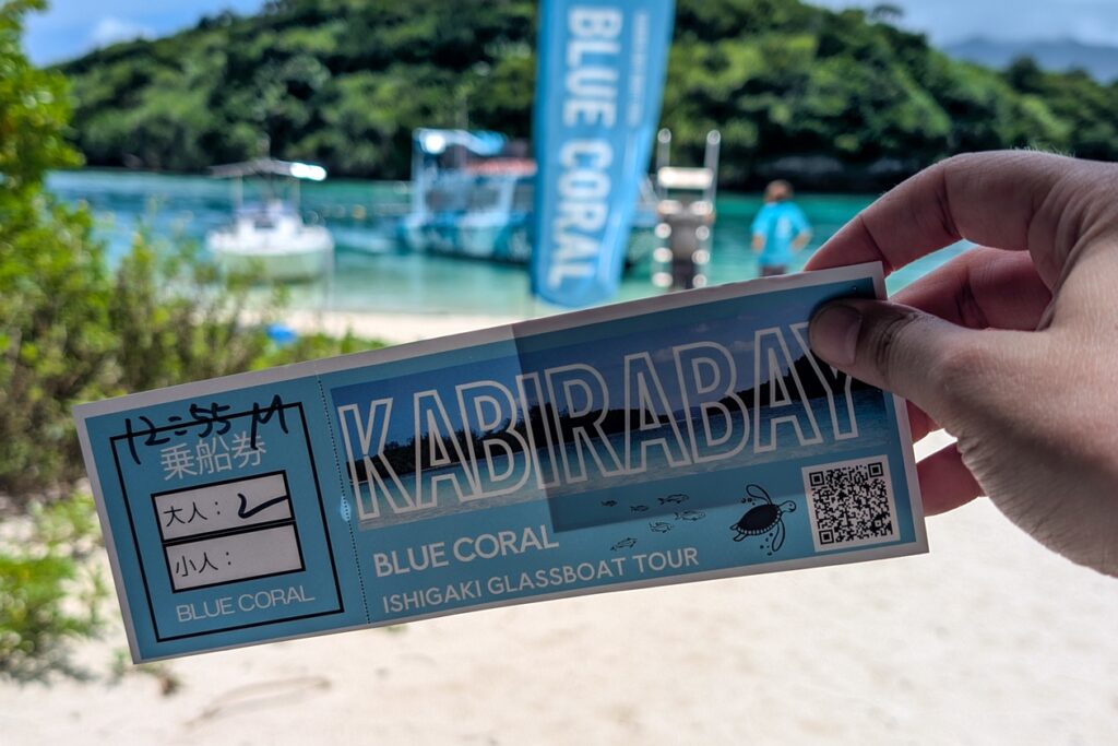 Eine Hand hält eine Eintrittskarte für eine Tour mit dem Glassbottom Boat in der Kabira Bay.