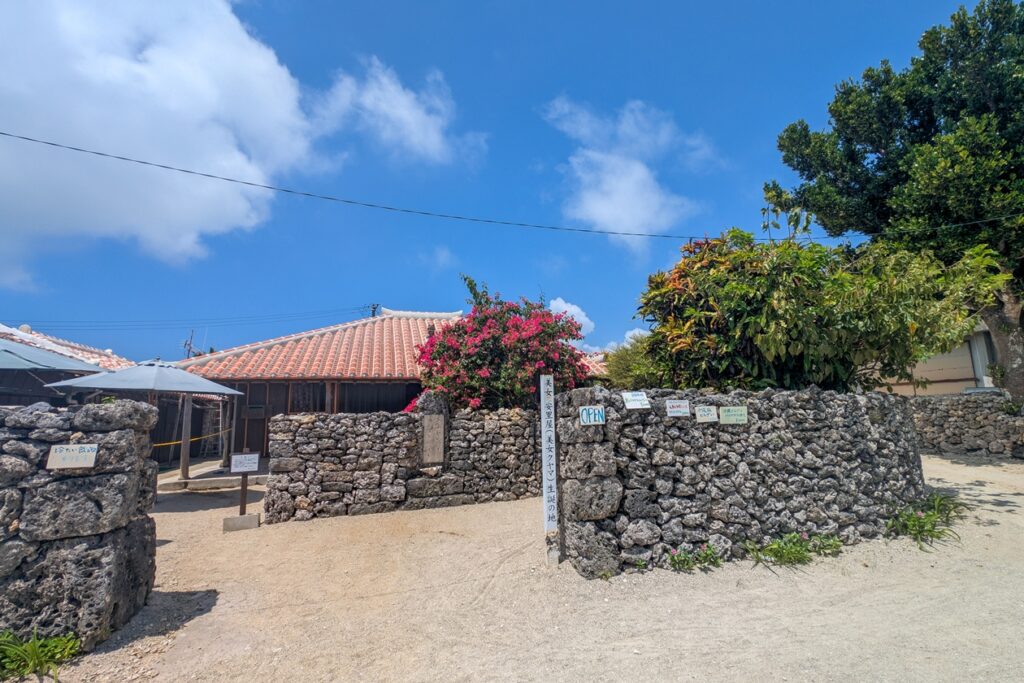 Traditionelle Architektur auf Taketomi. Das Haus hat ein rotes Ziegeldach auf welchem ein Shisa thront, Korallensandwege ziehen sich durch das Dorf und Gukku-Steinmauern säumen den Weg.