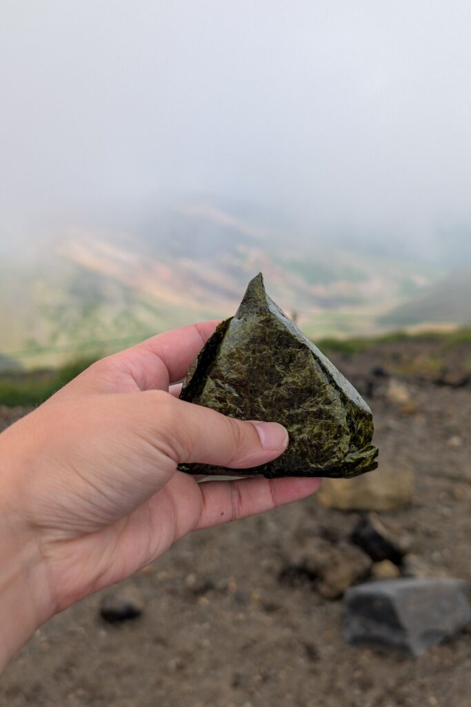 Eine Hand hält einen Onigiri. Im Hintergrund sind ein steiniger Untergrund und in der ferne bunte Berge zu sehen. Diese sind allerdings von Wolken bedeckt.
