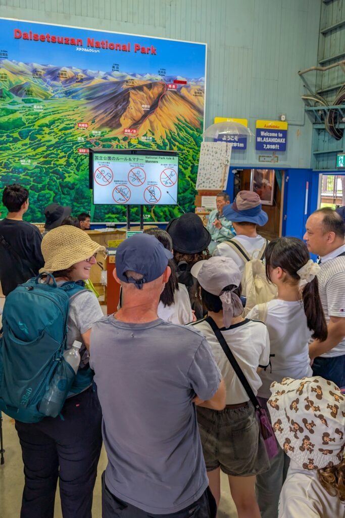 Einweisung durch einen Mitarbeiter des Daisetsuzan Nationalpark vor der Wanderung auf den Mt. Asahidake. Im Hintergrund ist eine Karte des Nationalparks zu sehen. Um den Mitarbeiter hat sich eine Traube aus Menschen gebildet.