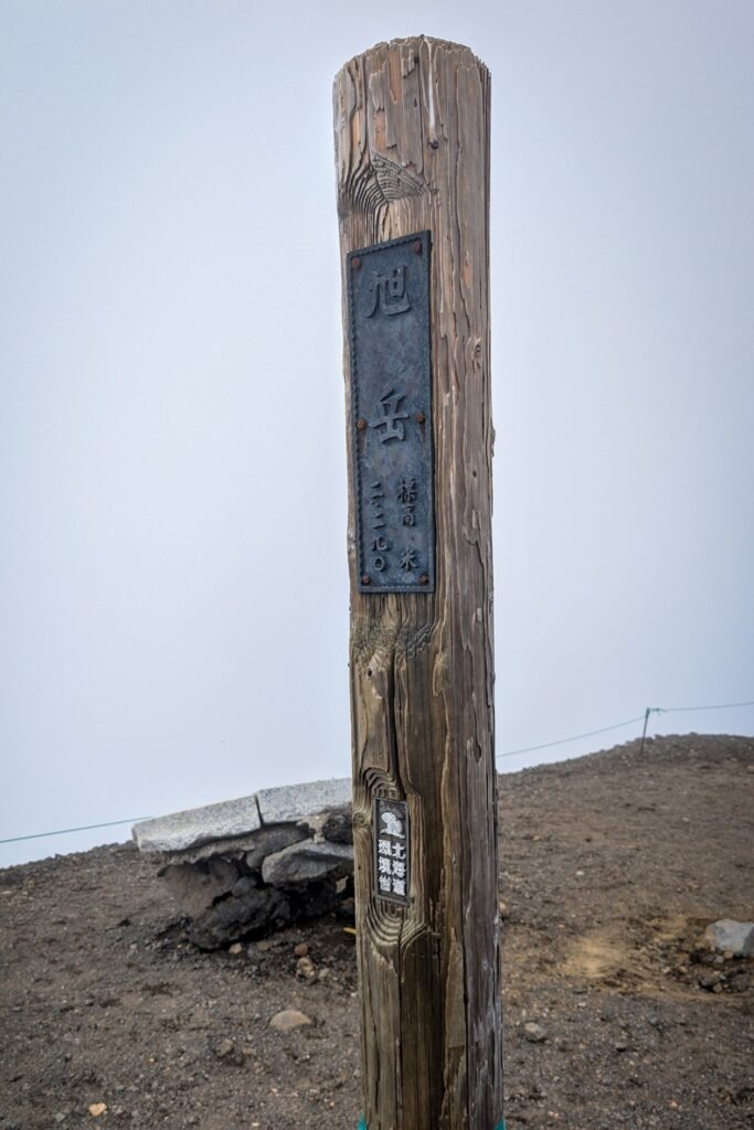 Holzpfahl mit japanischen Schriftzeichen, welcher den Gipfel des Mt. Asahidake markiert. 