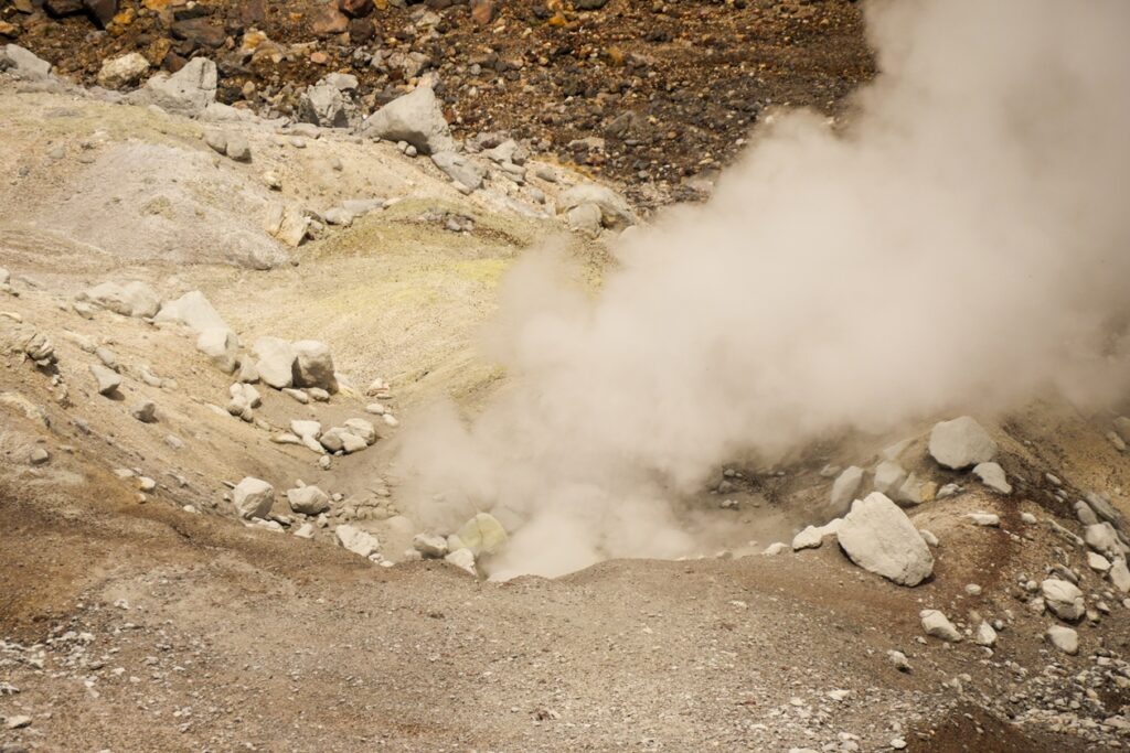 Detailaufnahme der Fumarole des Mt. Asahidake. Im Hintergrund sieht man weitere Berge des Mt. Asahidake. Das grün steht in einem starken Kontrast zu den erdtönen im Vordergrund.