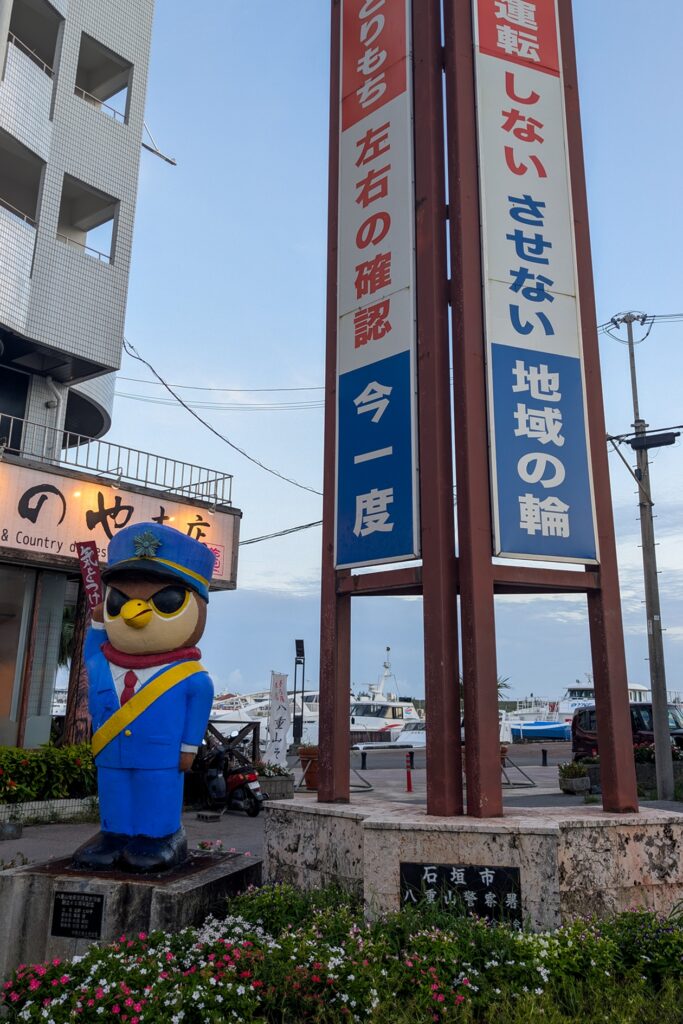 Der Vogel in Uniform und mit erhobenen rechten Flüge ist Maskottchen von Ishigaki und steht im Zentrum der Inselhauptstadt. Daneben ist eine Säule mit japanischen Schriftzeichen. Im Hintergrund sieht man den Hafen.