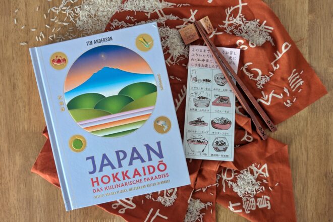 Das Kochbuch Japan, Hokkaidō das kulinarische Paradies liegt auf einem roten Tischläufer mit Schriftzeichen. Ebenfalls zu sehen sind Essstäbchen, ein japanischer Flyer und Reiskörner