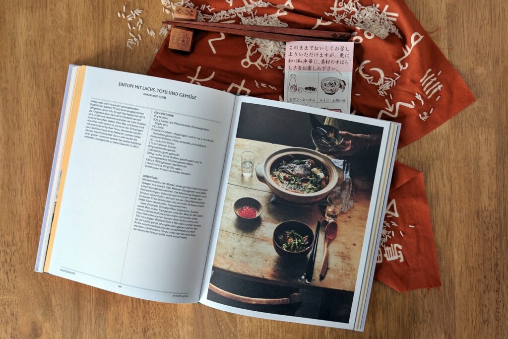 Blick ins das Kochbuch Japan, Hokkaidō - Das kulinarische Paradies. Es liegt auf einem roten Tischläufer mit Schriftzeichen. Ebenfalls zu sehen sind Essstäbchen, ein japanischer Flyer und Reiskörner