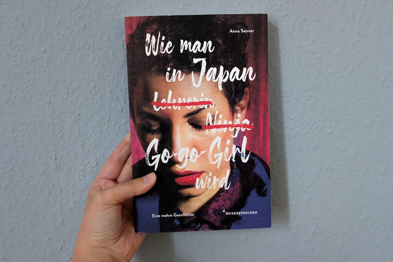 Eine Hand hält das Buch Wie man in Japan Go-go-Girl wird von Anne Sanner gegen eine hellblaue Wand.