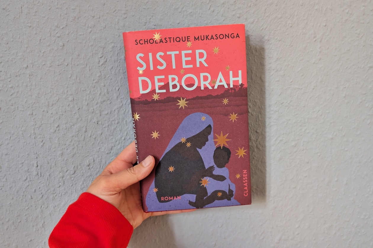 Eine Hand hält das Buch Sister Deborah von Scholastique Mukasong gegen eine hellblaue Wand