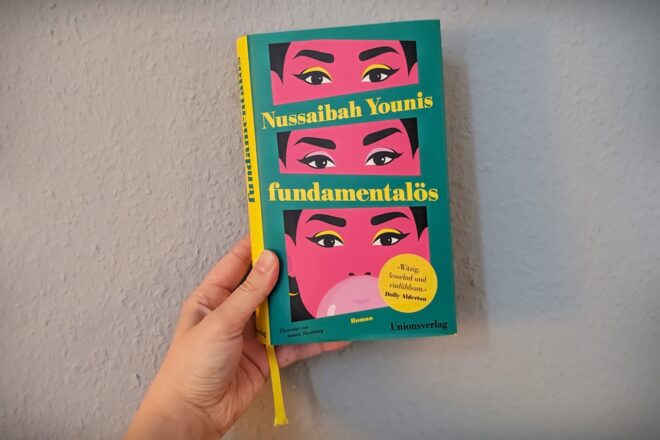 Eine Hand hält das Buch fundamentalös von Nussaibah Younis gegen eine hellblaue Wand.