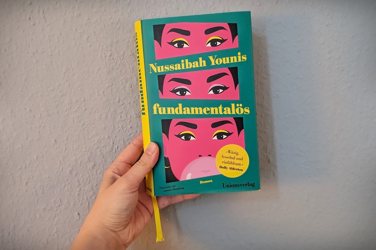 Eine Hand hält das Buch fundamentalös von Nussaibah Younis gegen eine hellblaue Wand.