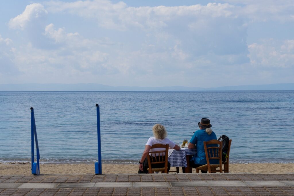 Zwei Personen sitzen an einem kleinen Tisch mit kariertem Tischtuch am Strand und blicken aufs Meer