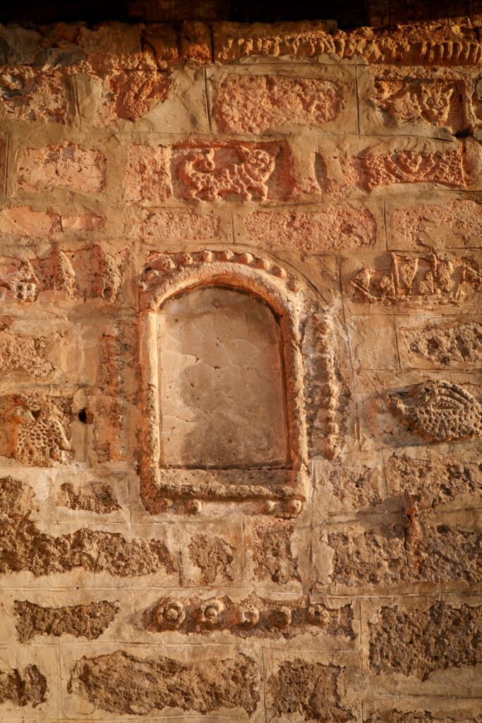 Mauer aus groben Steinen mit einem kleinen, zugemauerten Bogenfenster und dekorativen Reliefs
