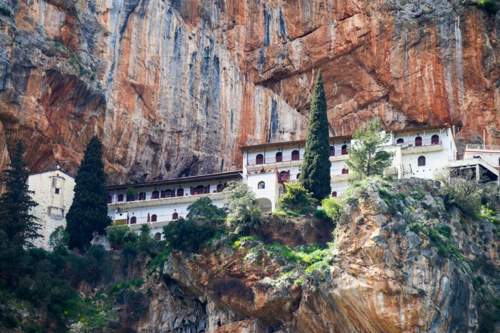 Gebäude des Klosters Elonis mit Arkaden und roten Dächern an einer steilen Felswand mit zwei hohen Zypressen davor