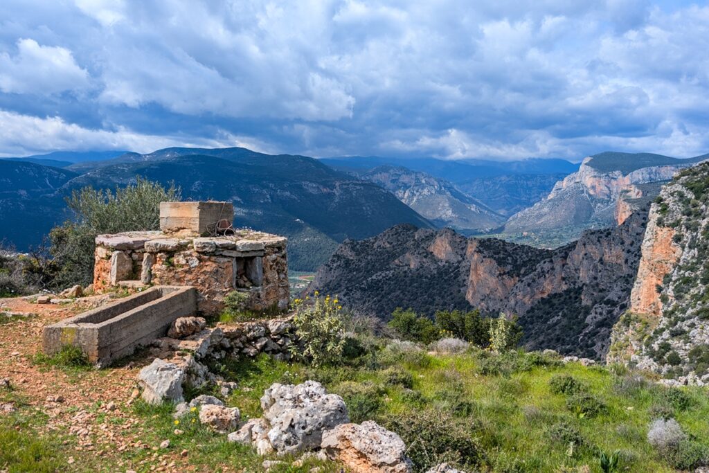 Alter Brunnen aus Stein auf einem Hügel mit Blick auf die Kletterfelsen rund um Leonidio unter bewölktem Himmel.