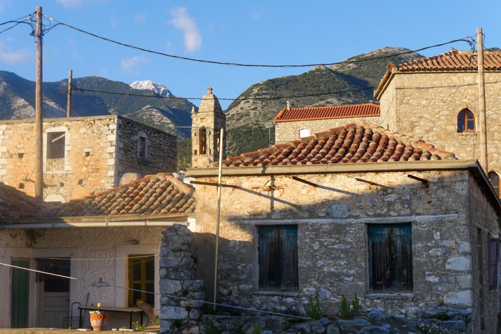 Steinbauten mit roten Ziegeldächern vor bewaldeten Bergen und blauem Himmel