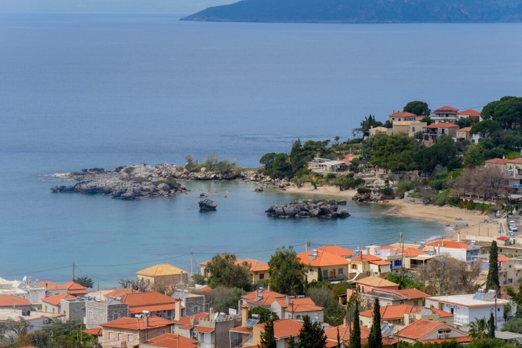 Bucht mit Blick auf den Ort Stoupa mit roten Dächern, Felsen im Wasser und bewaldeten Hügeln im Hintergrund