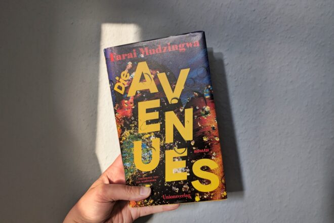 Eine Hand hält das Buch "Die Avenues" von Farai Mudzingwa gegen eine hellblaue Wand.
