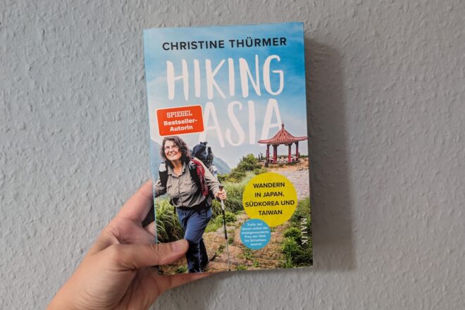 Hand hält Buch mit Titel 'Hiking Asia' von Christine Thürmer, auf dem Cover Wanderer mit Rucksack und asiatischer Pavillon im Hintergrund