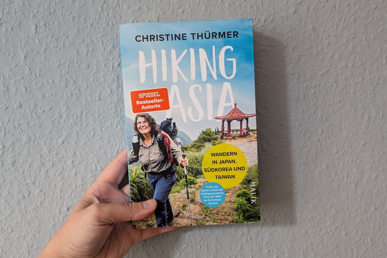 Hand hält Buch mit Titel 'Hiking Asia' von Christine Thürmer, auf dem Cover Wanderer mit Rucksack und asiatischer Pavillon im Hintergrund
