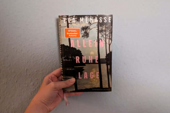 Eine Hand hält das Buch "Alleinruhelage" von Eva Menasse gegen eine hellblaue Wand.