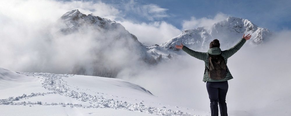 Lynn steht mit dem Rücken zur Kamera und reckt die Hände in den Himmel. Sie steht auf frischem Schnee und blickt auf zwei Berggipfel, die mit Schnee überzogen sind und von Wolken umspielt werden. Der Himmel ist blau.